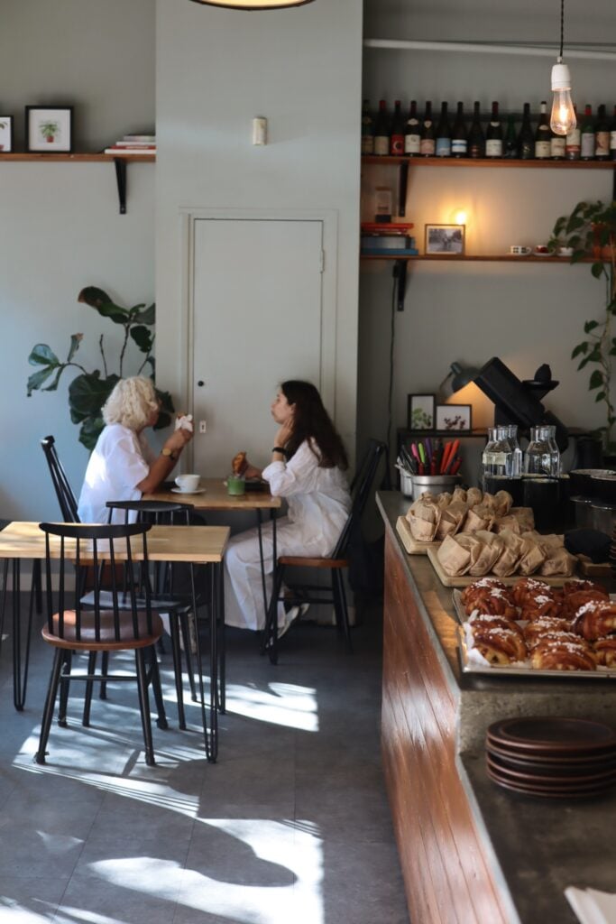 Två kvinnor, en med blont småkrulligt hår och en med svart hår, båda i vita kläder, sitter vid ett bord i ett café. Kanelbullar på bänken. 