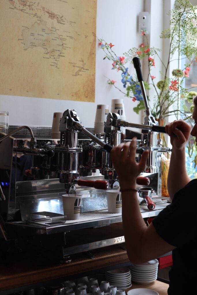 Två händer håller i handtagen på en avancerad kaffemaskin som häller upp kaffe i vita pappersmuggar. 