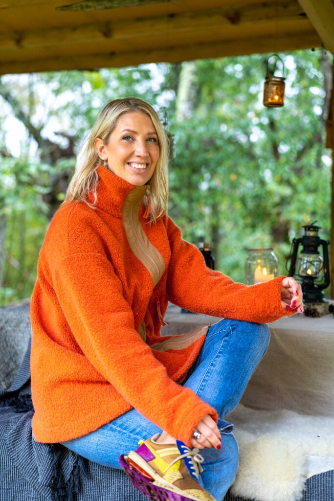 En blond kvinna i orange fleecetröja med blå byxor ler mot kameran, framför ett grönt buskage.