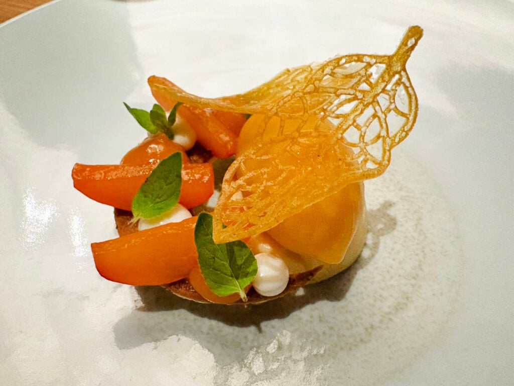 EN mycket elegant maträtt på vit tallrik, med orangea fruktklyftor och ett orange flarn i form av ett löv. 