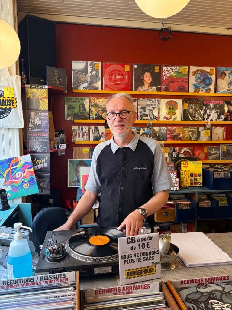 En man i piké, grått hår, glasögon, står bakom en LP-spelare i en butik med massa LP-skivor på väggarna. 