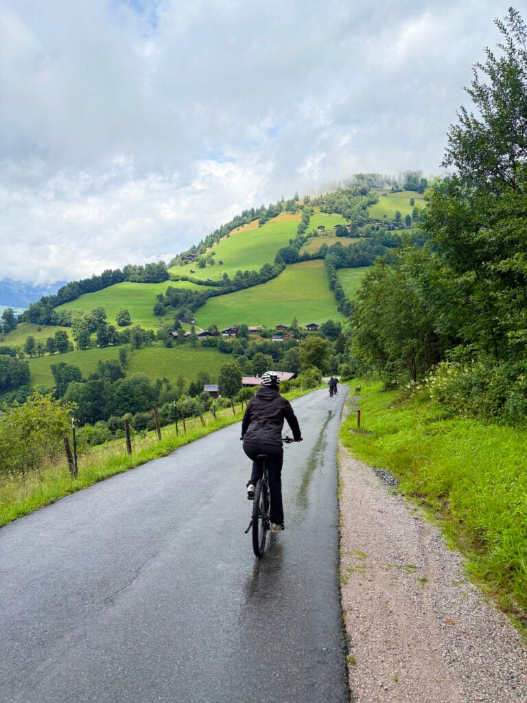 En blöt person i svarta regnkläder sitter på en cykel i ett böljande grönt bergslandskap. 
