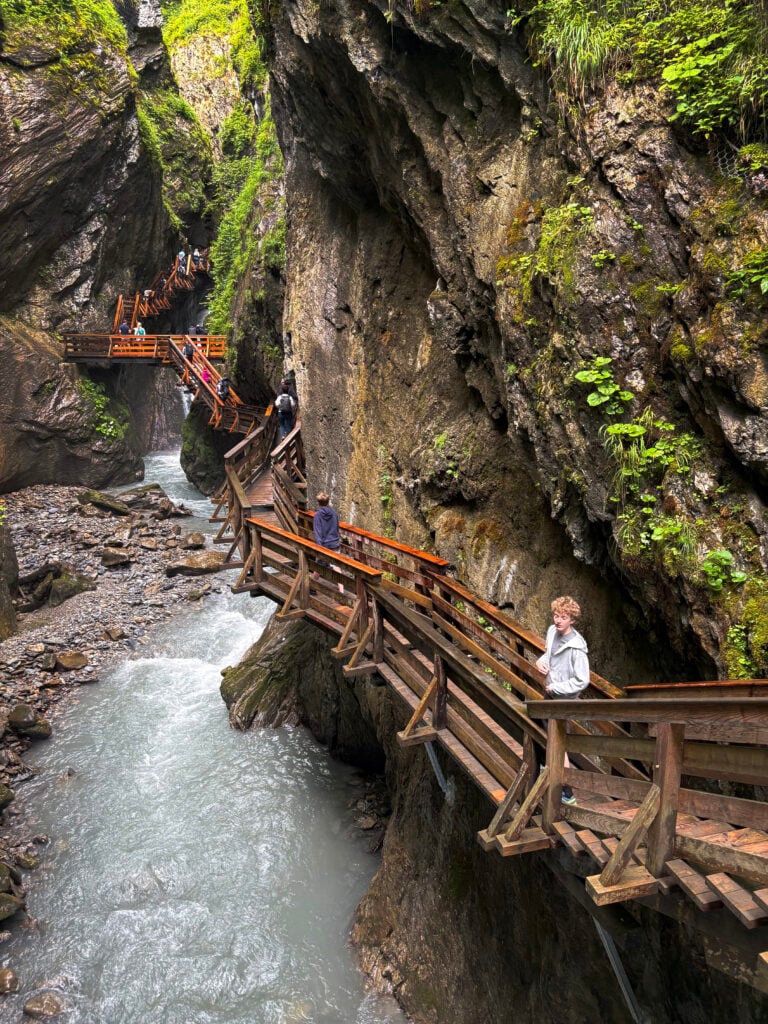 En träbro löper genom en trång ravin, en fors rinner bredvid, en person i vit tröja tittar på vattnet. 