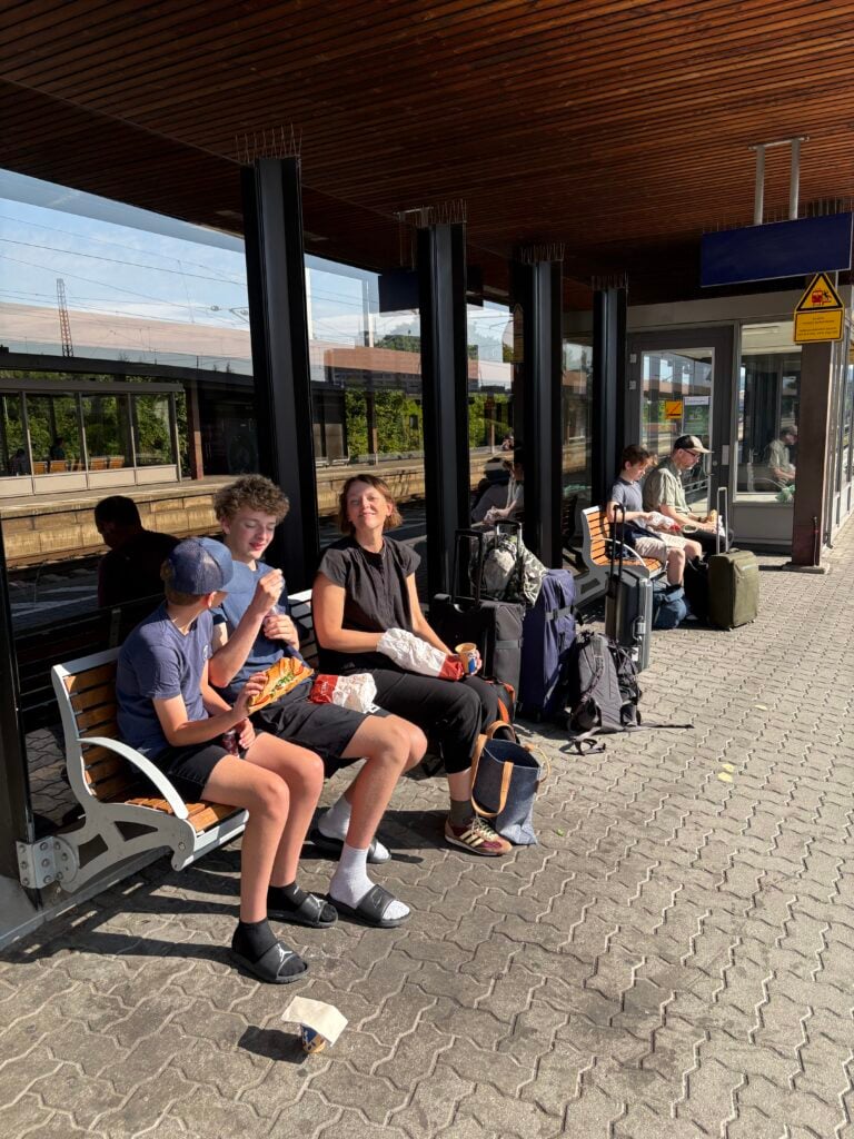 Flera vuxna och två tonåringar sitter på en rad bänkar på en tågstation i morgonsolen. 