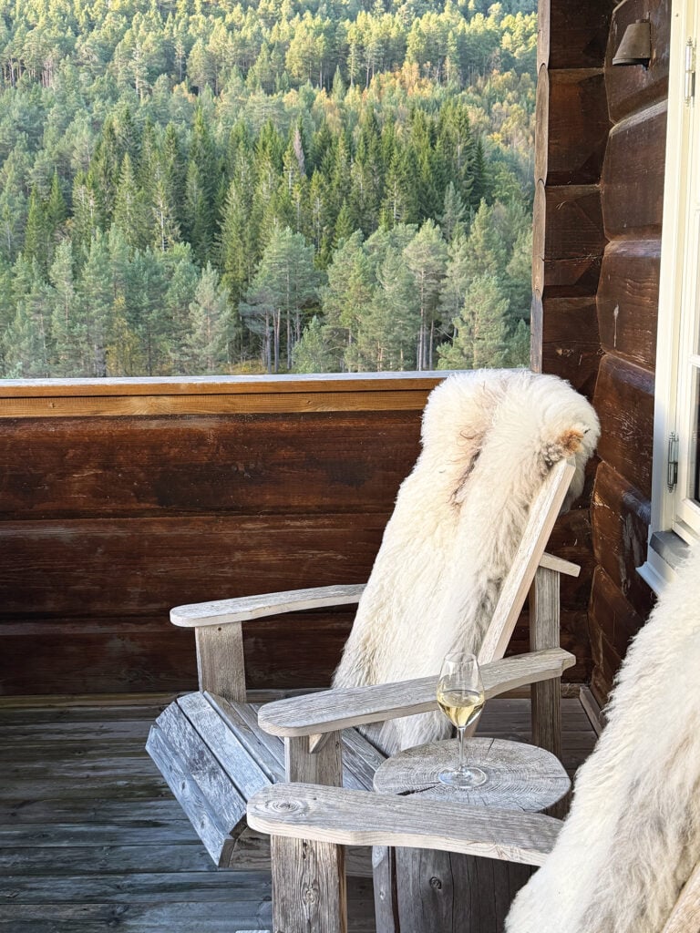 En rå trästol med fårskinn står på en timrad uteplats med skog bakom. 