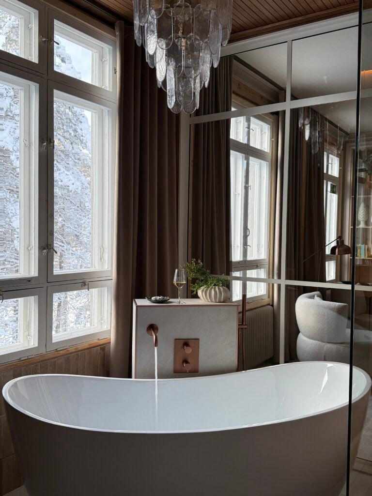 Ett rundat exklusivt badkar står i hörnet bredvid ett fönster med snötäckta träd utanför. 