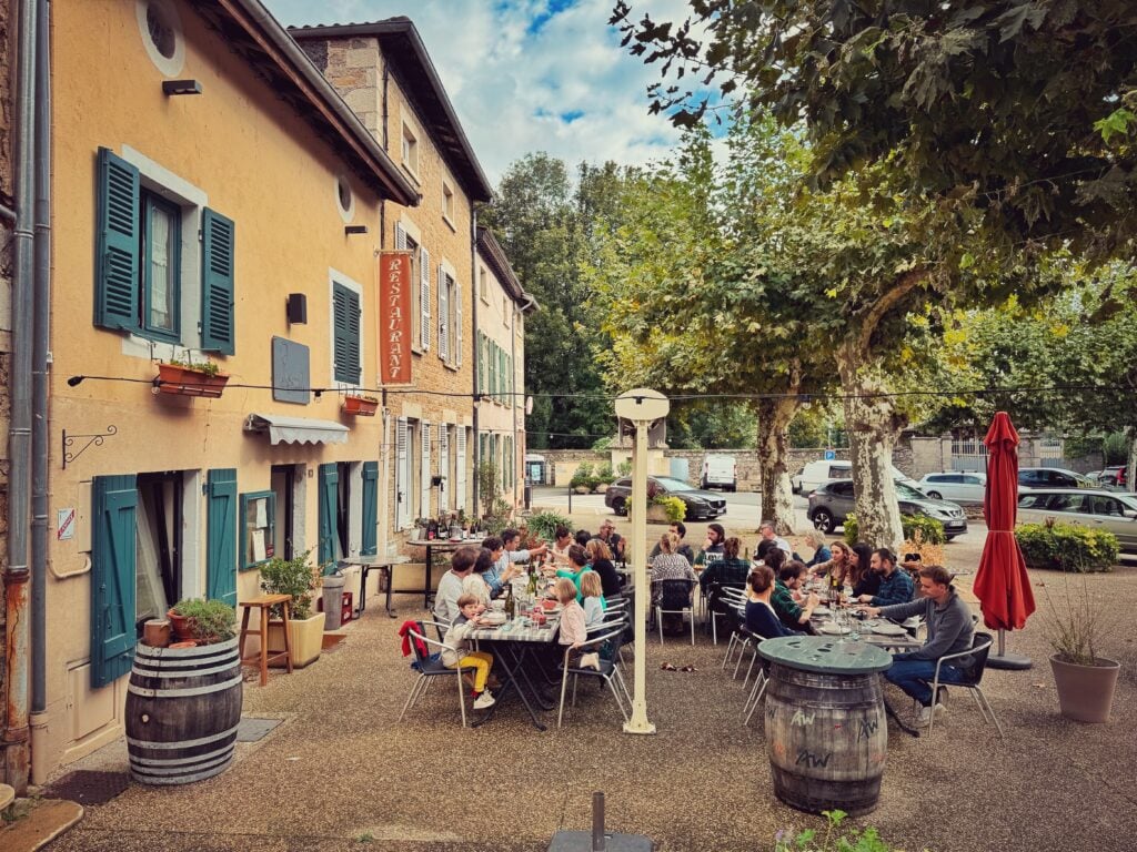 En mysig uteservering framför franska gula hus med fönsterluckor, lummiga träd. 