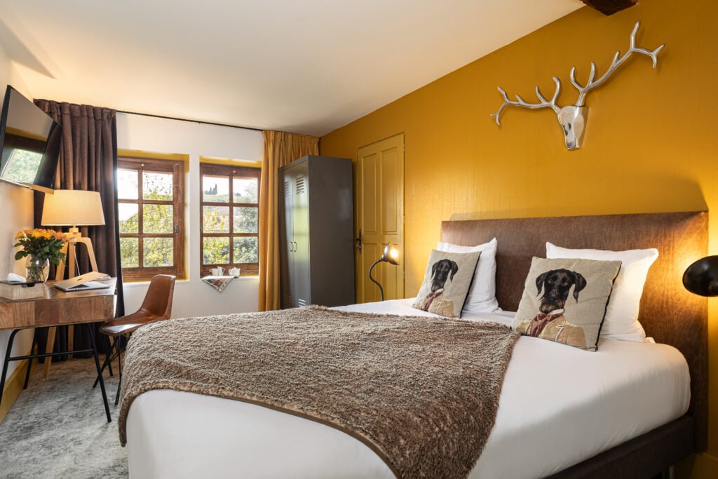 Ett hotellrum med gula väggar, en hjortskalle på väggen, sängen är bäddad med vita lakan, brun filt och hundar på kuddarna. 