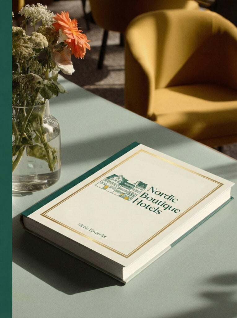 En bok med vitt omslag och grön text ligger på ett blekgrönt skrivbord med en blomvas bredvid.