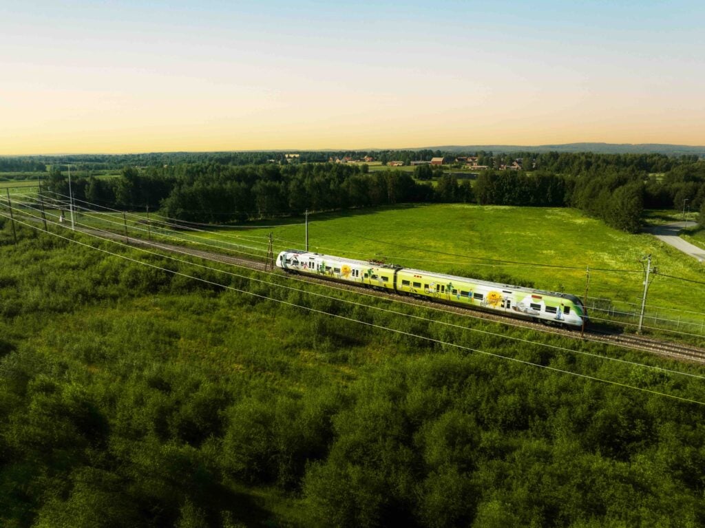 Ett vit-gult tåg susar genom gröna fält och skogar.