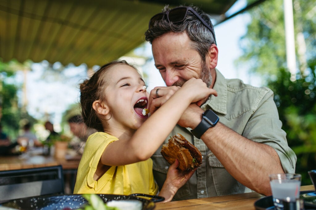 Cute girl having dinner with her father. Eating hamburger in restaurant, enjoying quality tasty food together. Delicious. Father's Day.