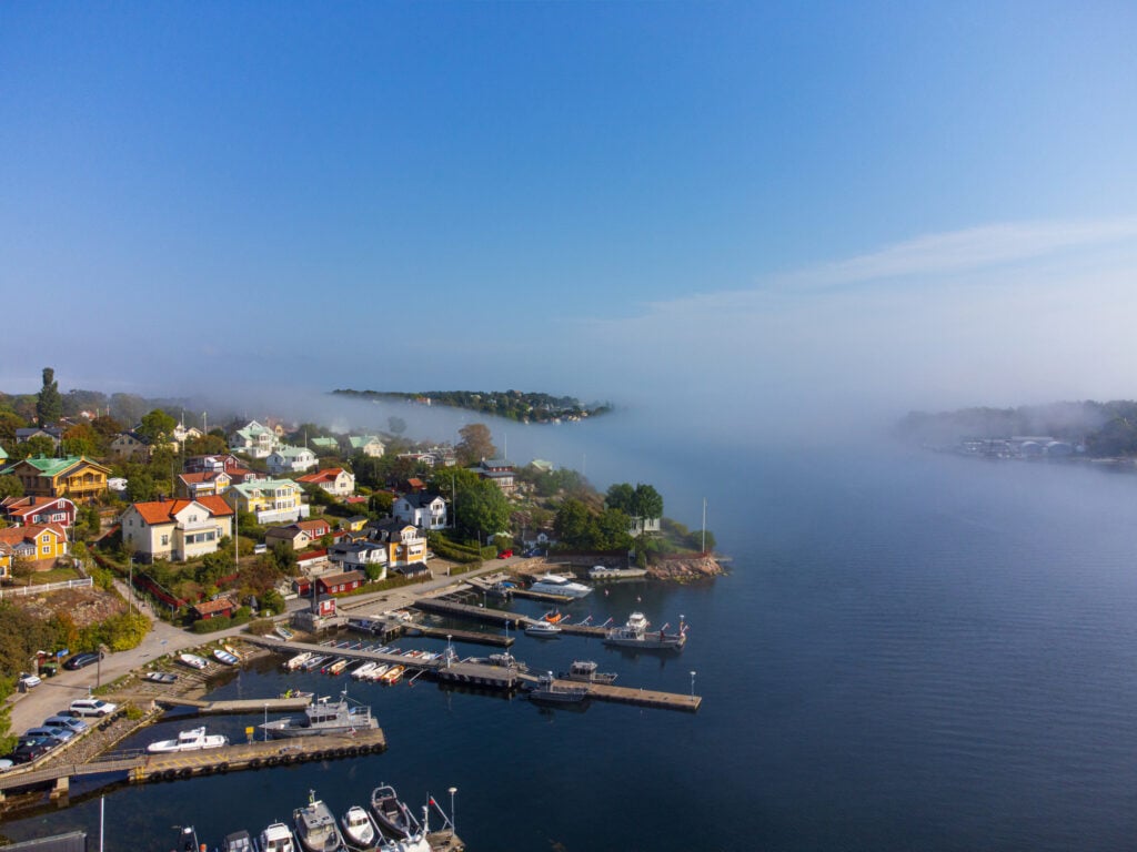 Liten by med bryggor vid havet, blå hav och himmel, en dimbank rullar in. 