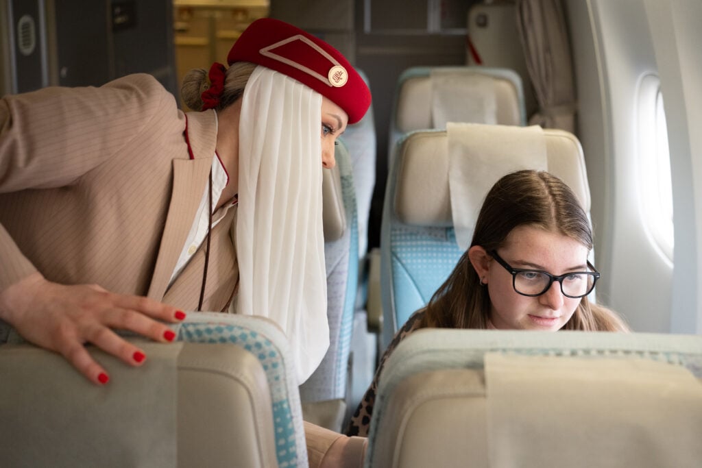 En flicka sitter i en flygplanssol, en flygvärdinna med röd hatt och vit sjal lutar sig över.