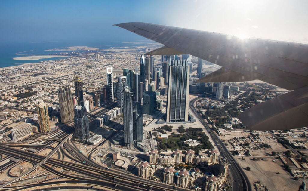En flygplansvinge skymmer sikten av Dubai från ovan. Skyskrapor och blått hav syns.