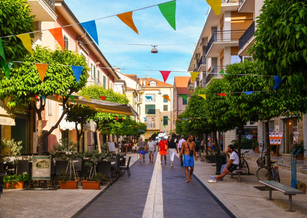 En liten italiensk by, gågata, små stadshus, människor som strosar, blå himmel. 