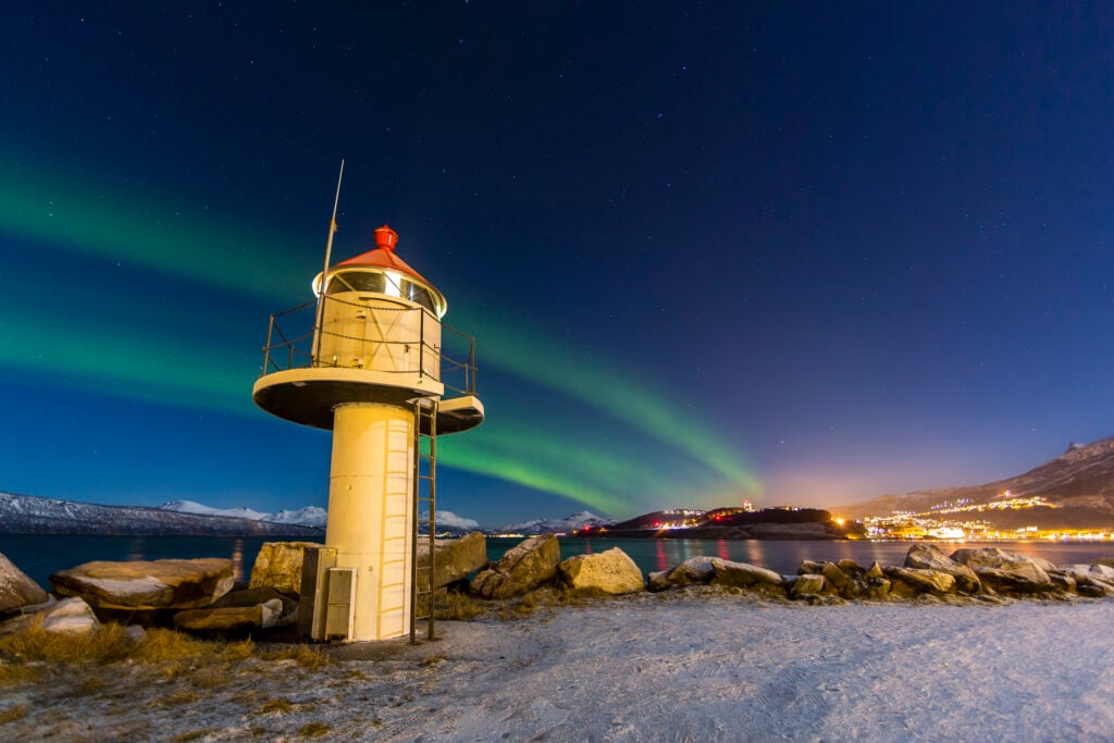 Norrskenet flammar över en upplyst fyr, lite snö på marken.