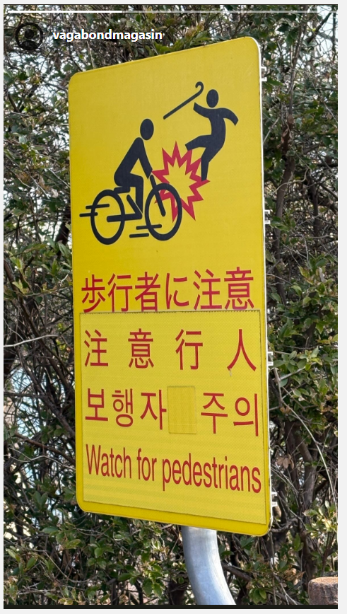 En gul skylt med japansk text och en bild på en cyklist som kör på en människa med käpp. 