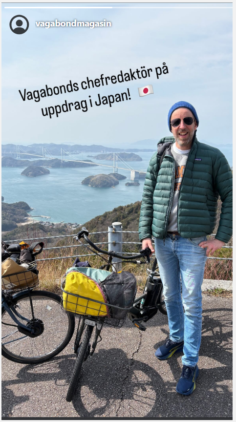 En man i grön tunn dunjacka står bredvid en cykel på en hög höjd, havet och små öar syns bakokm. 