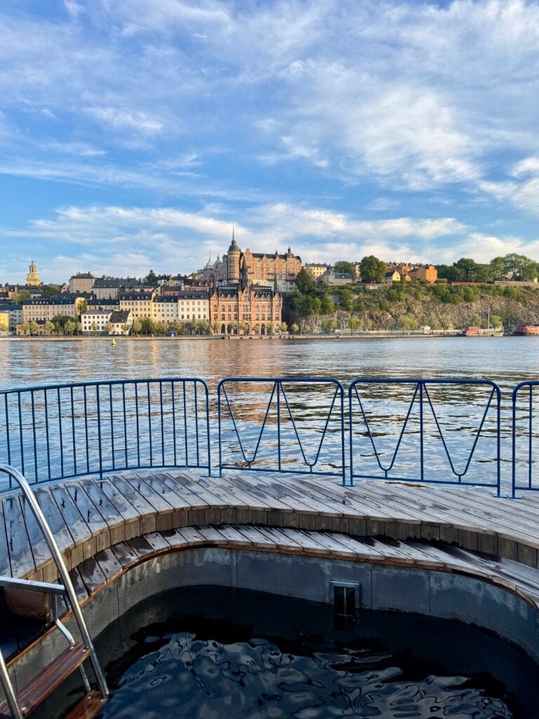 En rundad badbrygga omgärdas av staket, vattnet kluckar utanför. Münchenbryggeriet syns på Söders höjder.