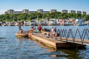 En brygga sträcker sig ut i vattnet med en rund badplattform längst ut, flera människor njuter av solen, en del har badkläder.