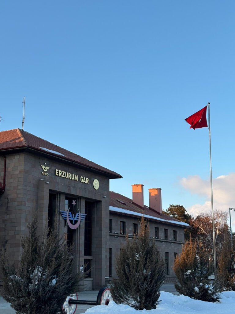 En grå stationsbyggnad med en röd flagga på stång, snö på marken, blå himmel. 