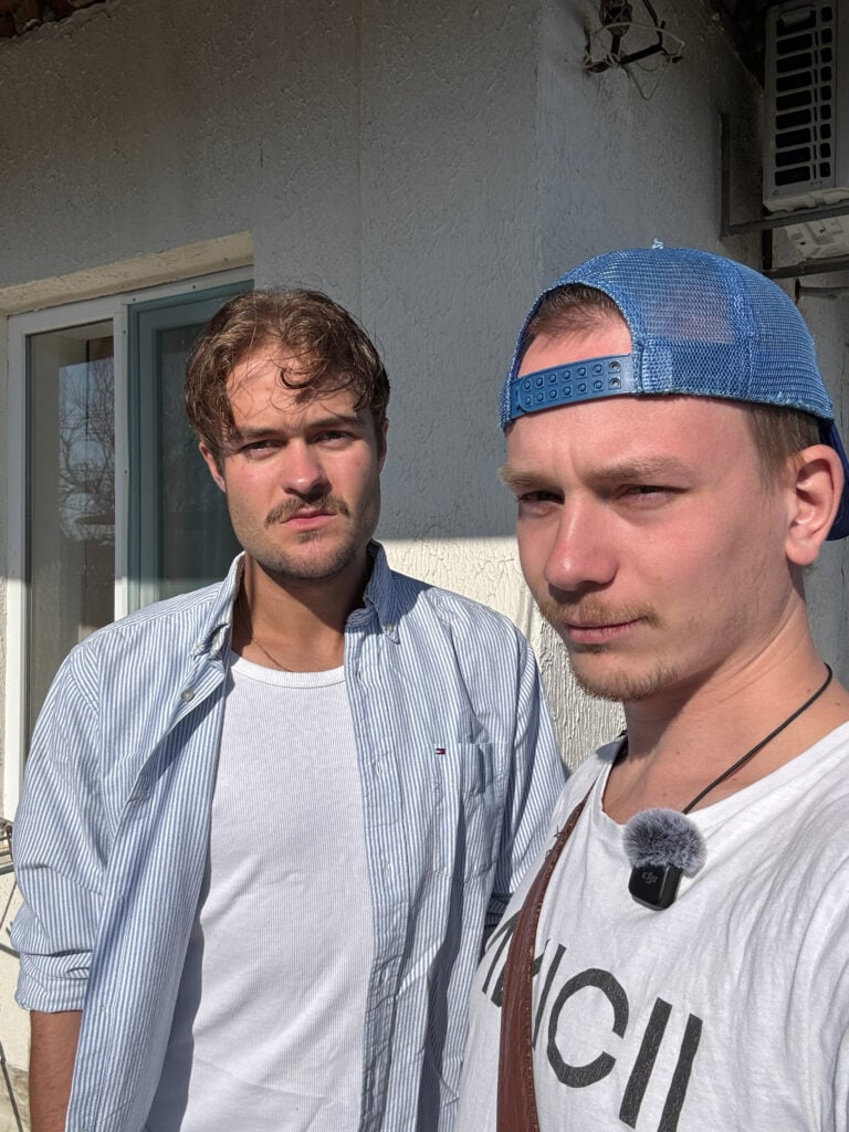 Två killar poserar mot kameran i solen, de ena har yvigt hår och ljusblå skjorta, de andra har blå keps bak och fram och en smal mustasch. 