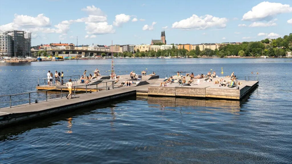 En brygga med stor badplats löper ut i vattnet, på andra sidan vattnet syns en stadssilhuett.