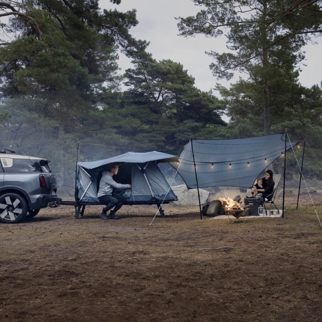 En bil med ett tält och ett par med en bebis som sitter under en tarp. Skog bakom. 
