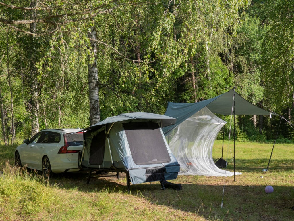 Ett tält bredvid en vit bil i en skog, en tarp med ett myggnät under. 