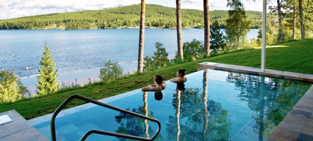 Två personer badar i en turkos pool med utsikt över skogar och en sjö. 