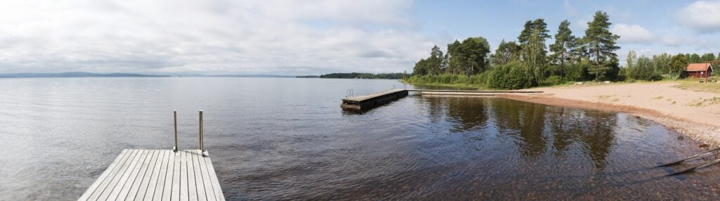 Panoramabild av en badbrygga och en udde som sticker ut i en sjö. 