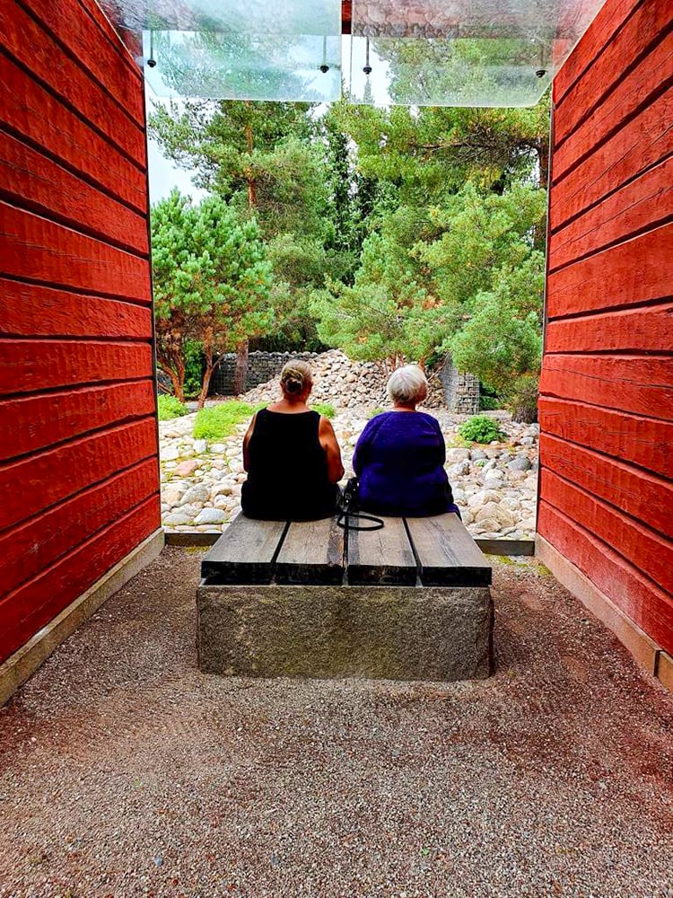 Två personer sitter i skuggan på en bänk mellan två röda husväggar, och tittar på en grönskande trädgård. Vy bakifrån. 