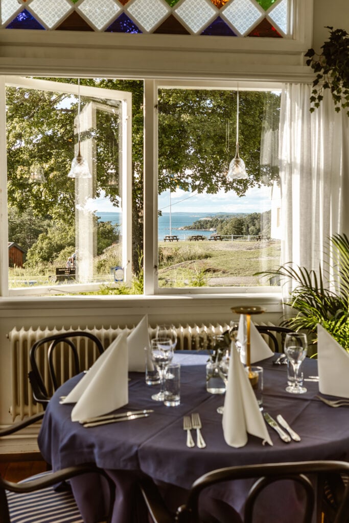 Ett dukat bord med blå duk och vita servetter, ett fönster avslöjar en utsikt mot grönskan och havet. 