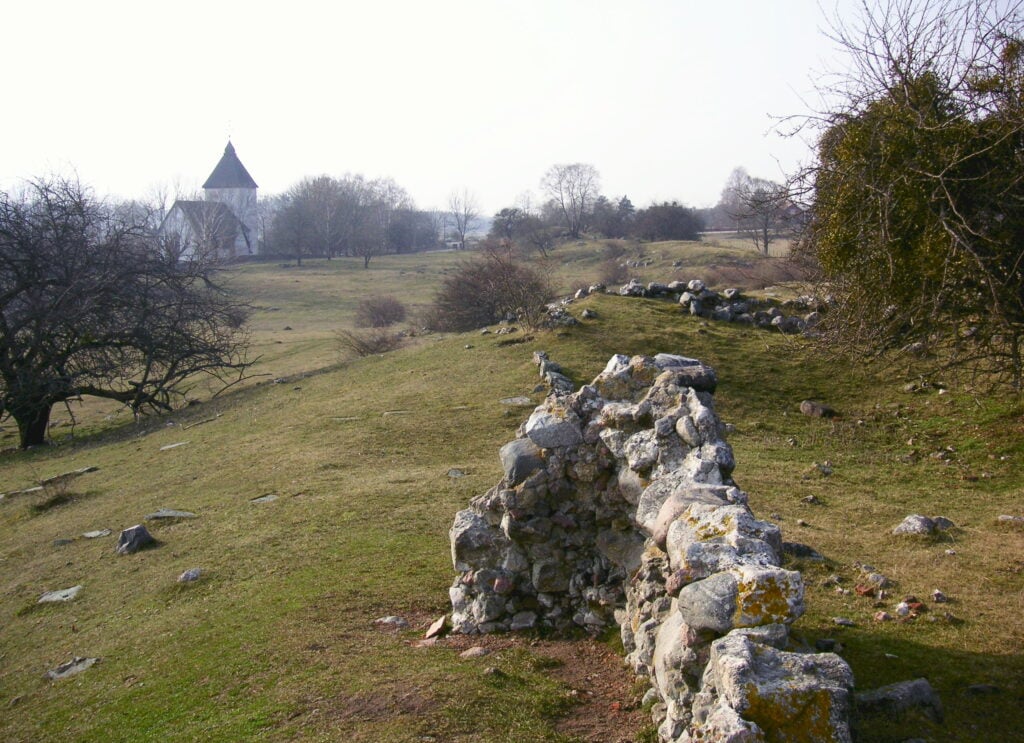 Stenruiner i gräslandskap med kyrka i bakgrunden.