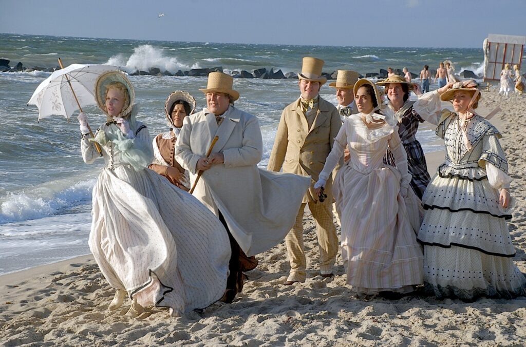 Människor i vita 1800-talskläder går på havsstrand med solparasoll och hattar.