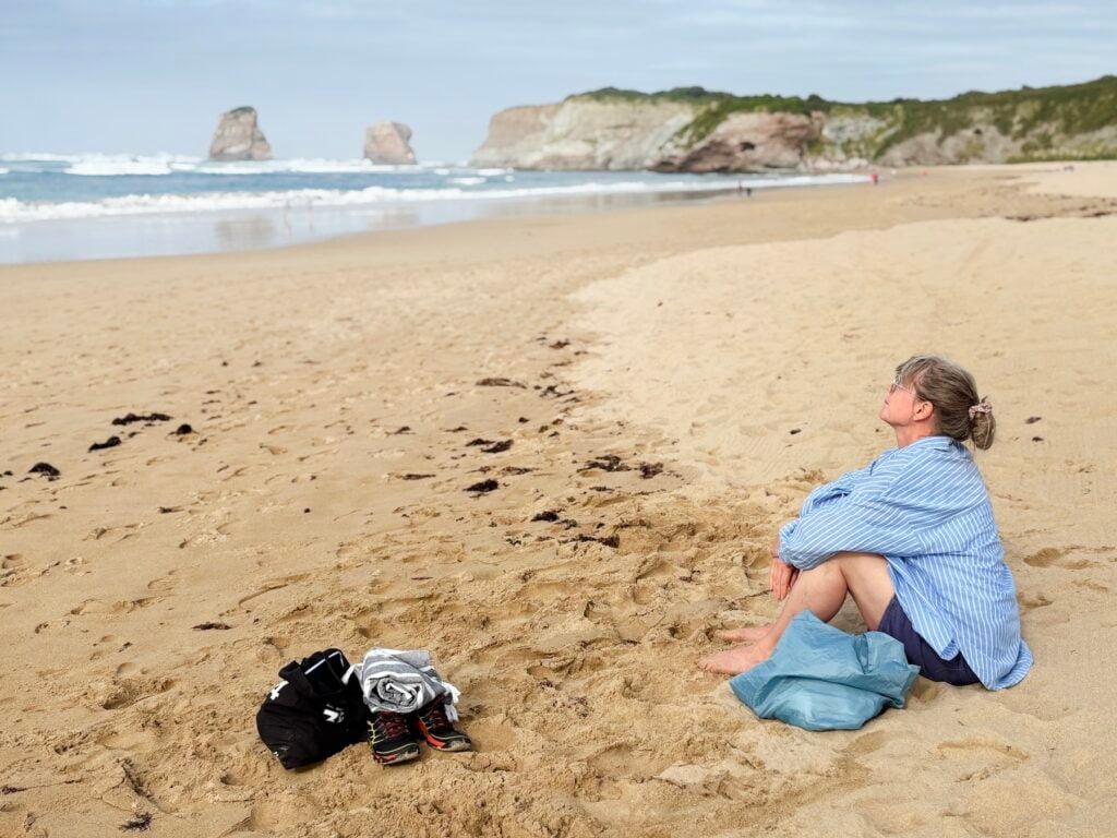 Kvinna i blå-vit skjorta sitter på sandstrand vid havet.