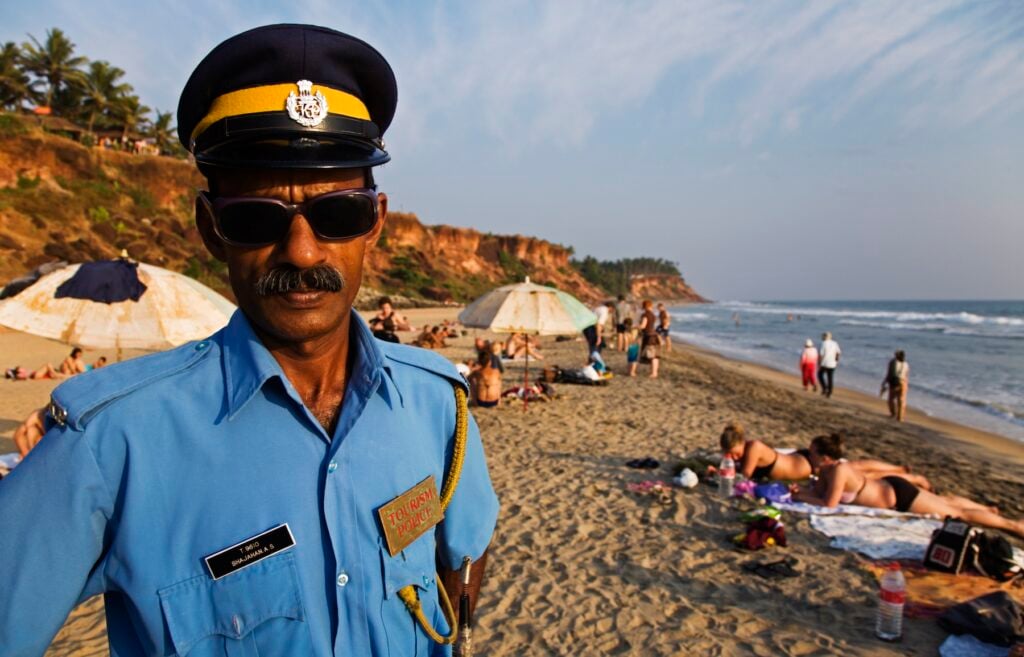 Polis i solglasögon och blå skjorta på sandstrand med solbadare.