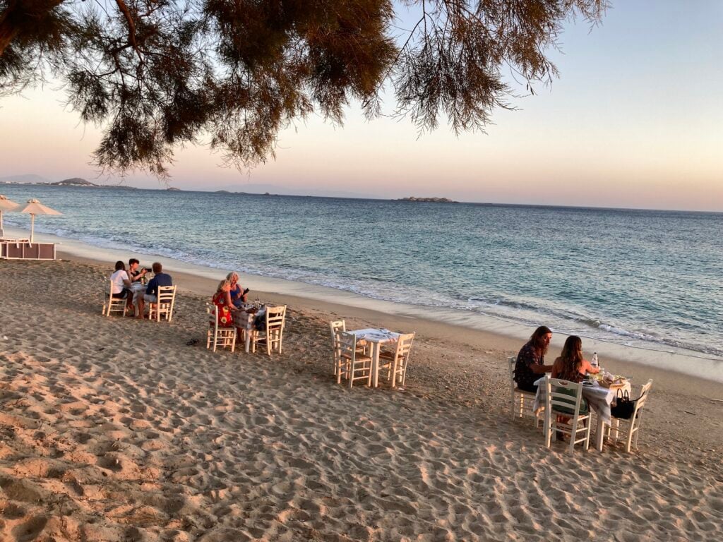 Strand med vita restaurangbord i sanden och hav i bakgrunden.