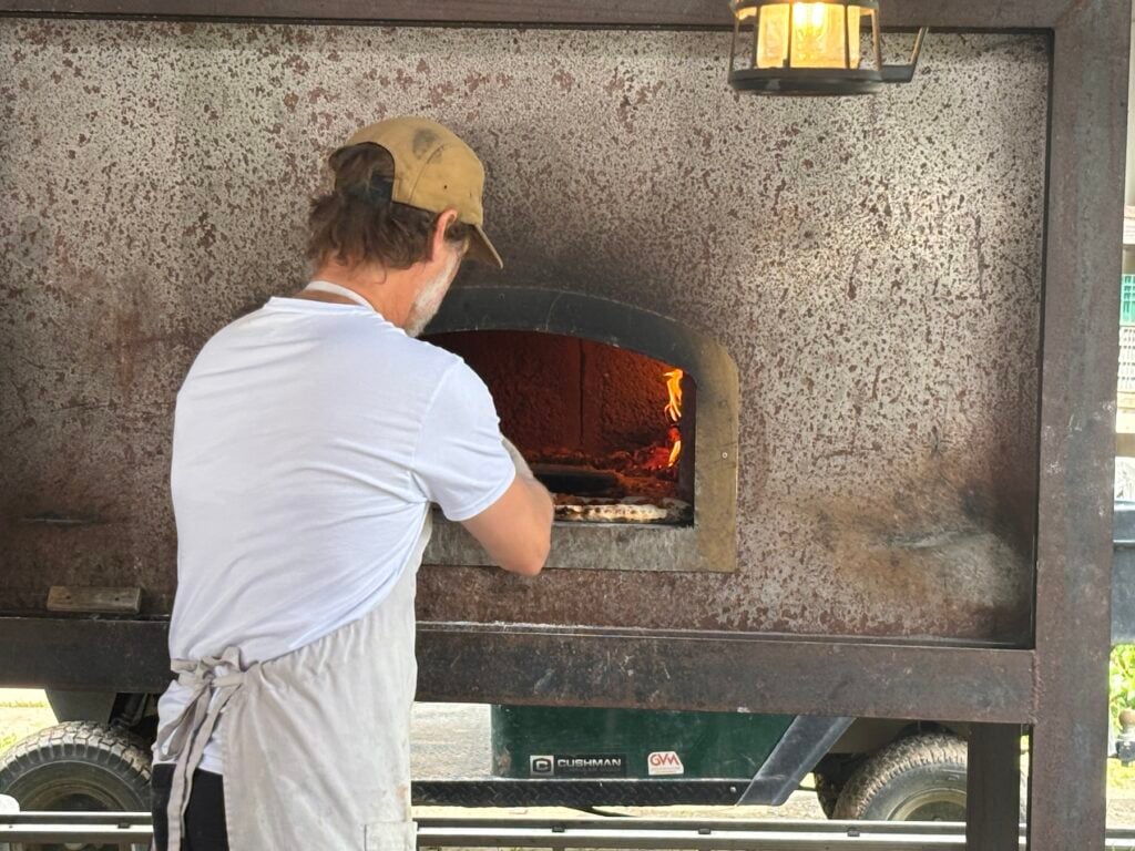 Man med keps som stoppar in pizza i vedeldad pizzaugn.