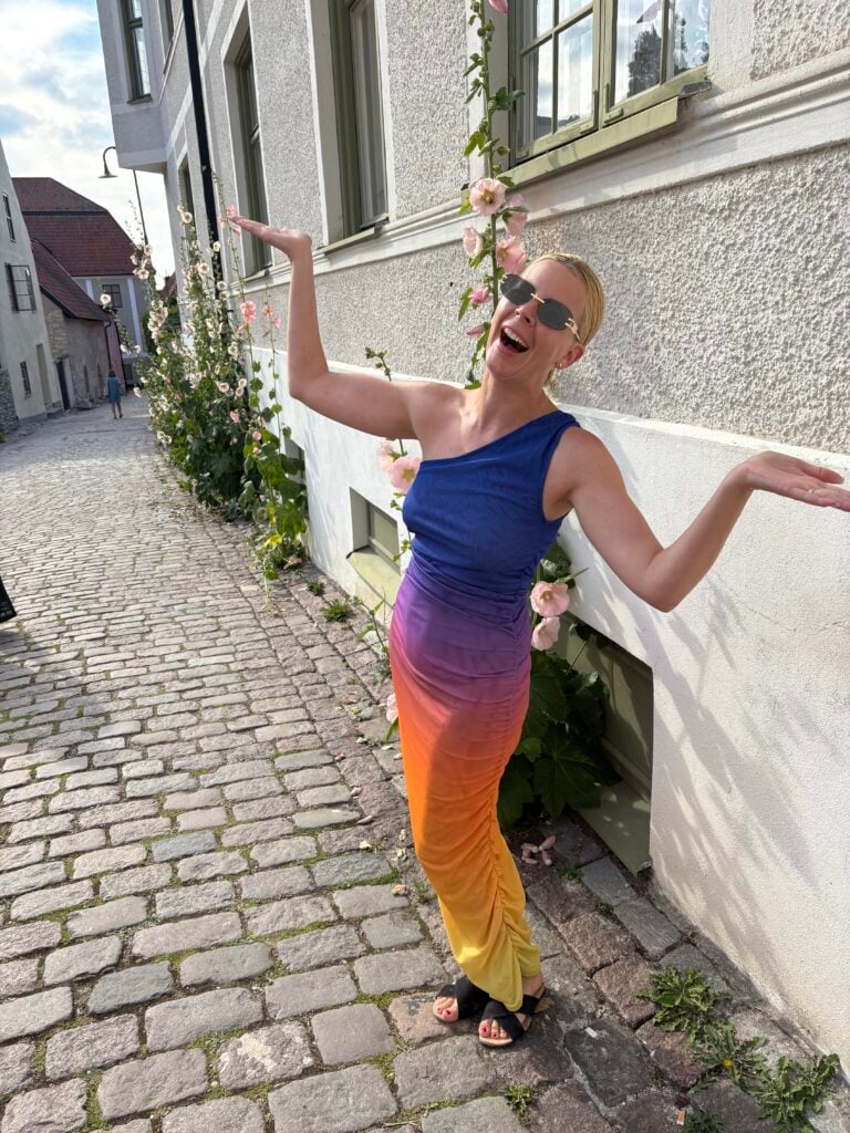 En kvinna i tight blå-lila-orange-gul klänning tramsar och poserar mot en kamera på en kullerstensgata.