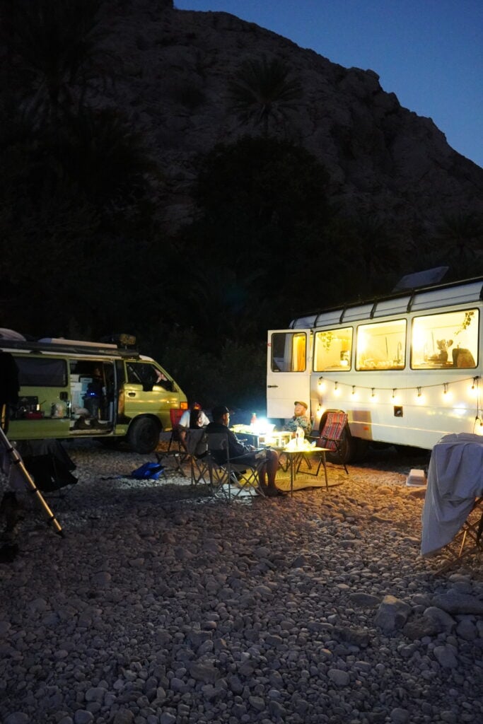 Två campingbilar, upplysta med ljusslingor, står parkerade i mörkret på en stenig strand.