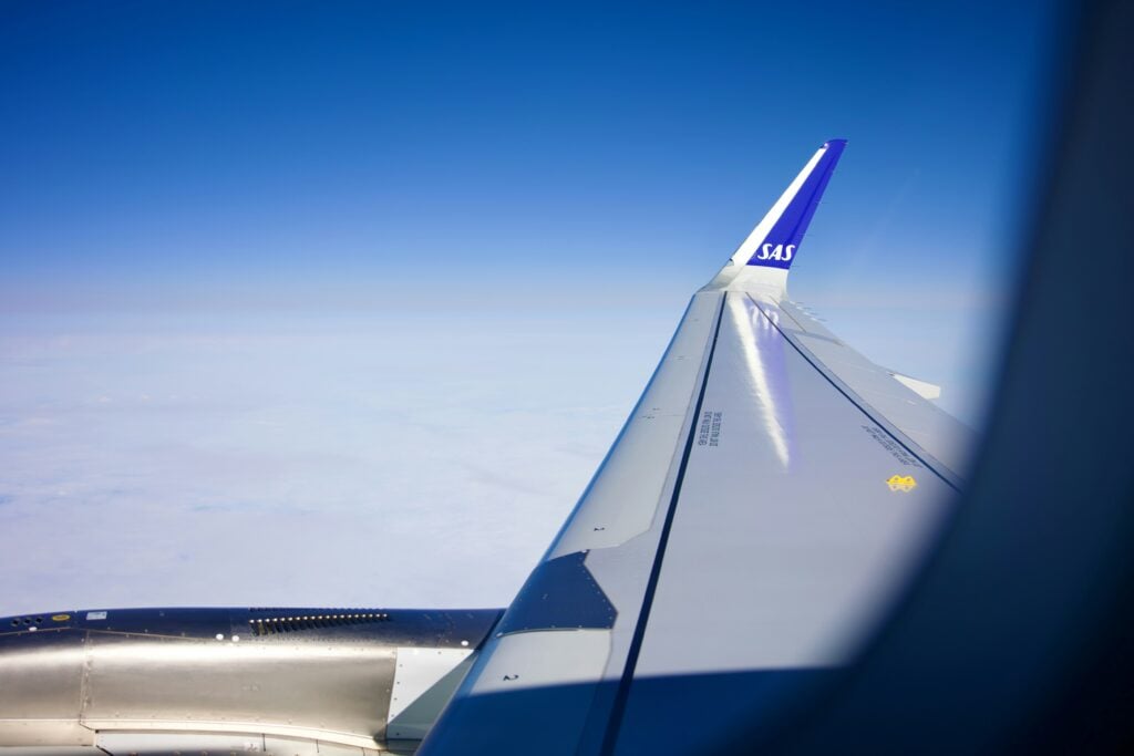 En flygplansvinge syns genom flygplansfönstret, på flygtur ovanför molnen.