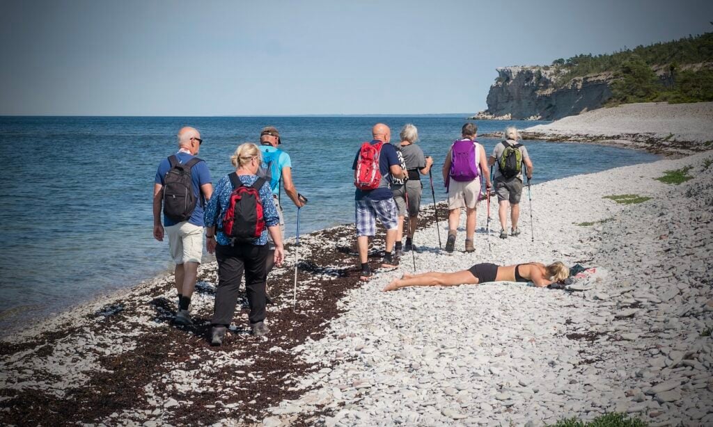 Sju äldre människor med ryggsäckar vandrar på vit stenstrand. En yngre kvinna ligger ner på stranden och solar.