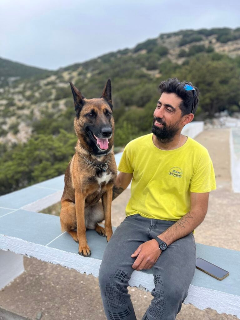 Man i gul t-shirt med hund sitter på bänk med berg och hav i fonden.