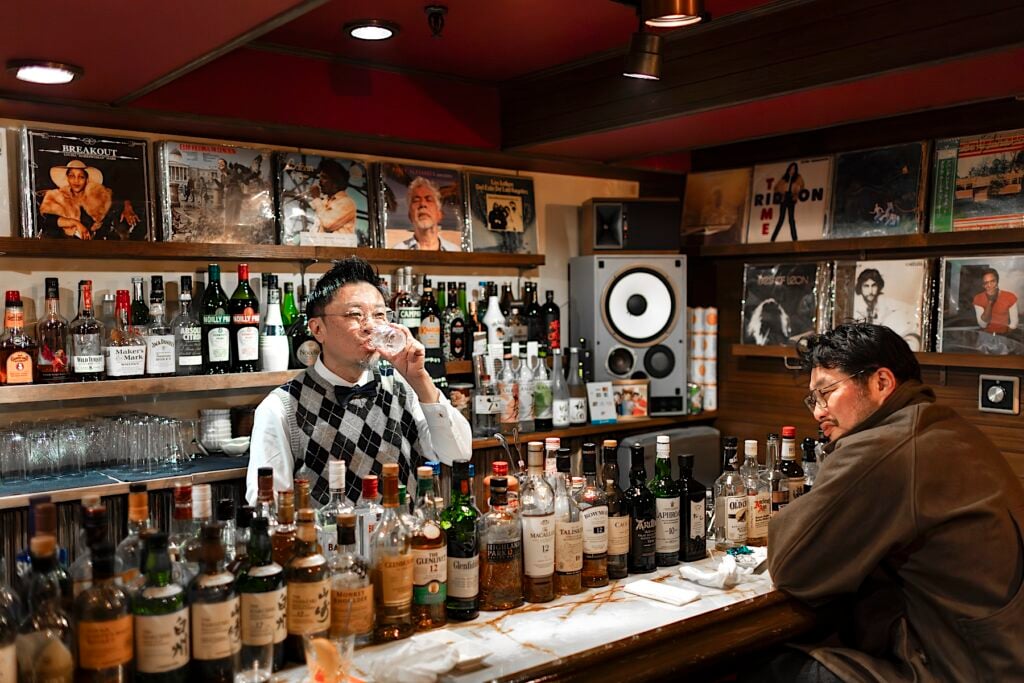 Bardisk med en bartender och en bargäst. Många flaskor på hyllorna.