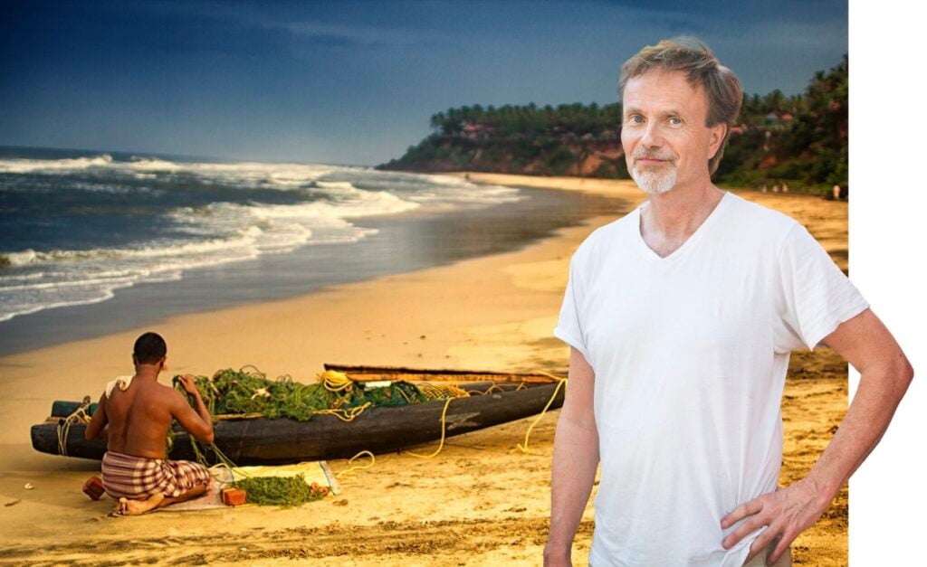 Man i vit t-shirt framför strand där fiskare sitt vid båt med fiskenät.