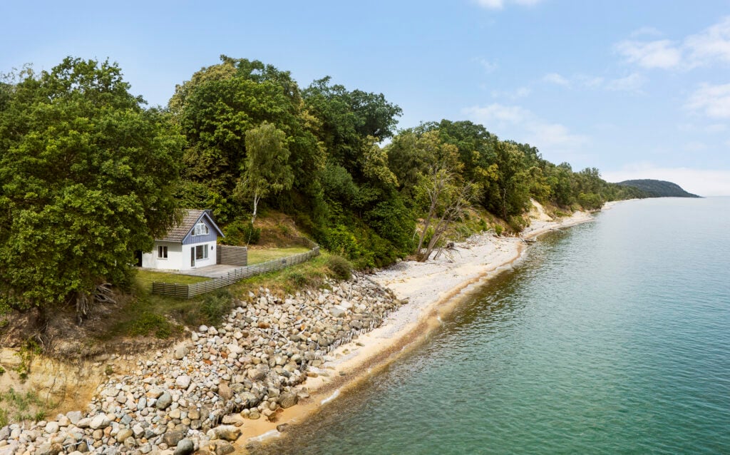 I en grön lövskog gömmer sig ett hus, på bredvid en vit sandstrand och ett lugnt hav.