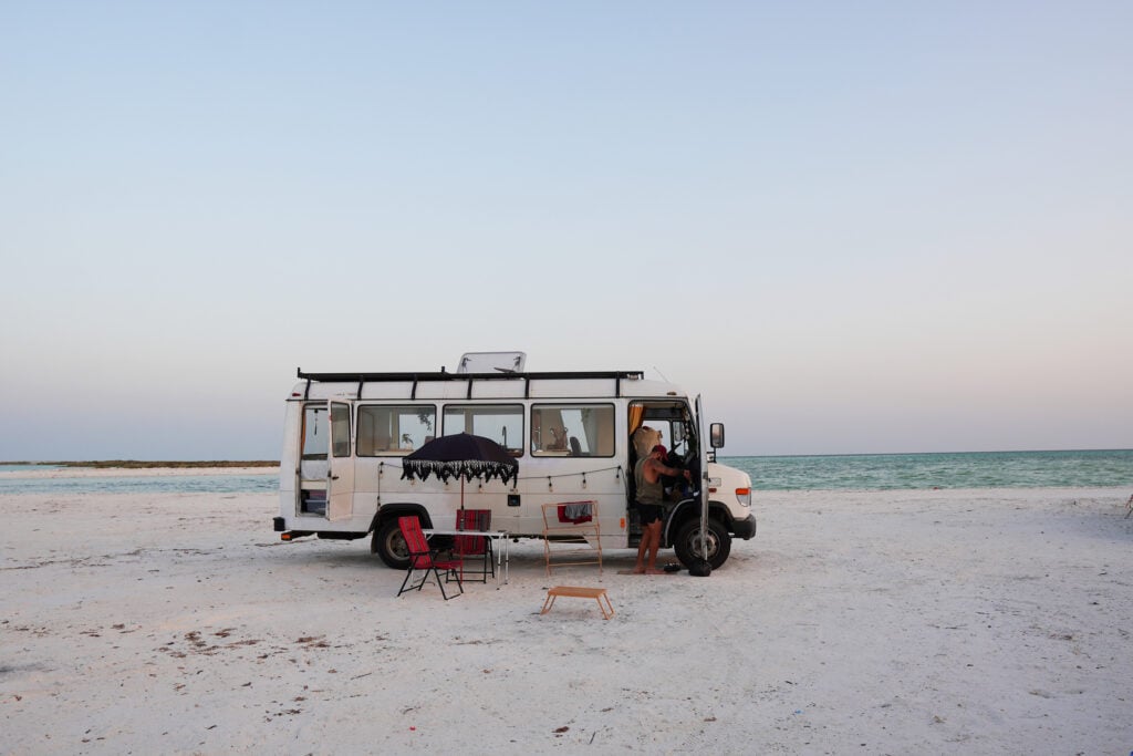 En vit campingbuss står parkerad på en kritvit sand framför ett turkost hav, pastellfärgad himmel.