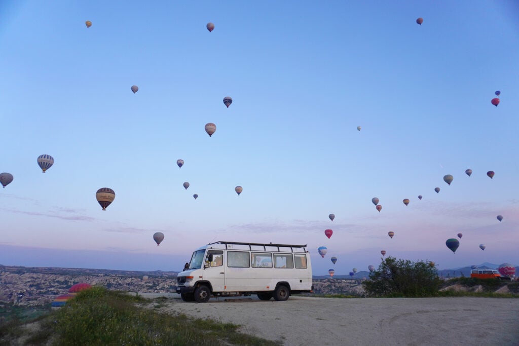 En vit buss står parkerad framför en höjd, pastellfärgad himmel, hundratals luftballonger flyger över.