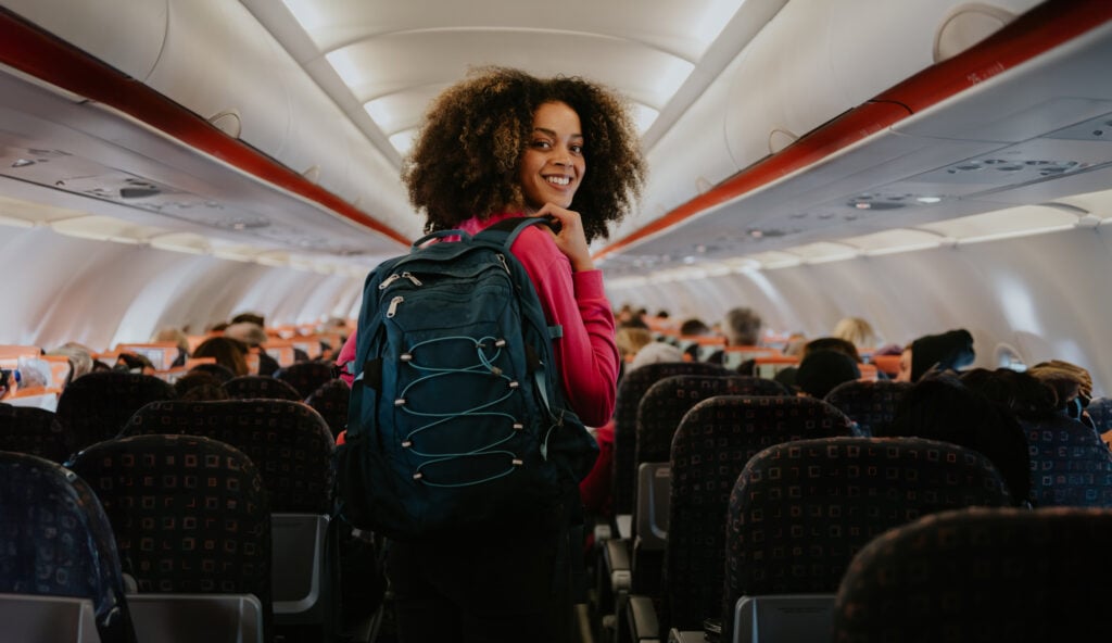 En mörkhårig ung kvinna går genom ett flygplan, ryggsäck, tittar bakåt och ler mot kameran.